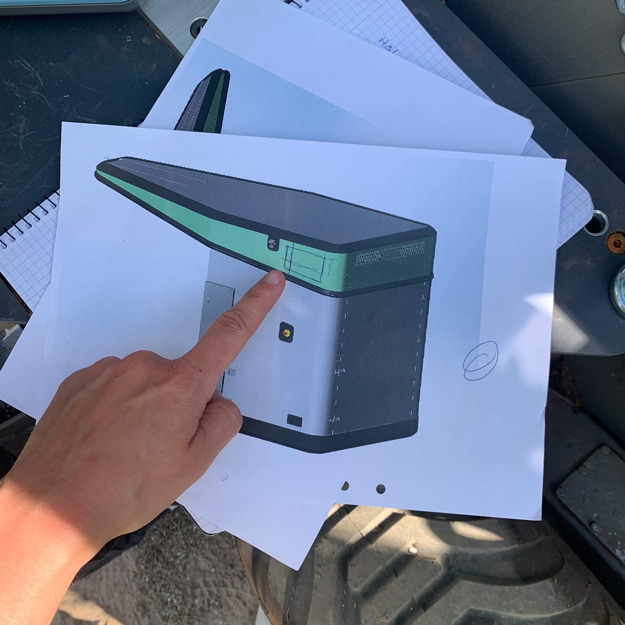 Finger that points at a printed sketch of the planting robot that lies on the physical robot's tire.
