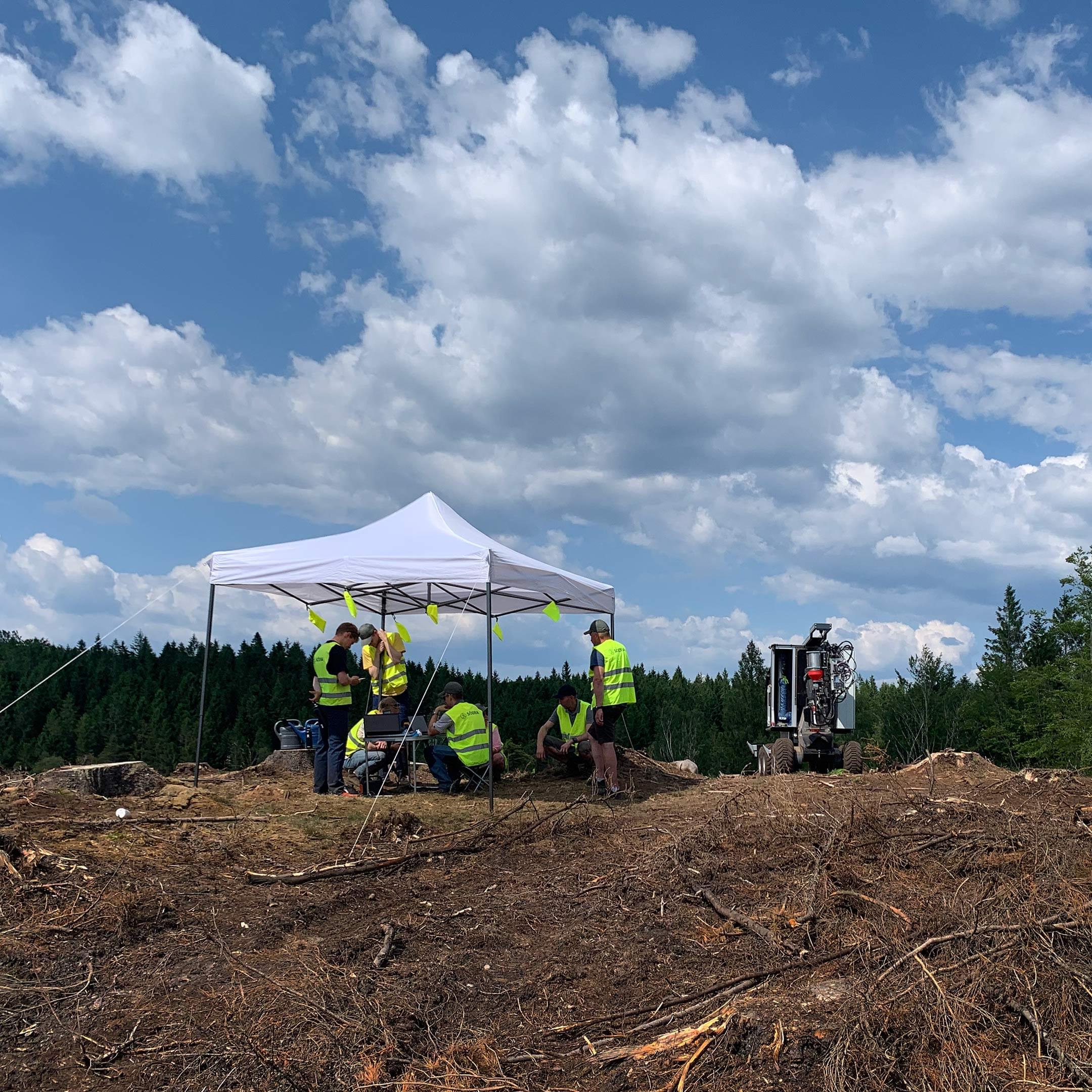 Five people standing under a sun tent in the forest. To the right of the tent stands a planting robot.